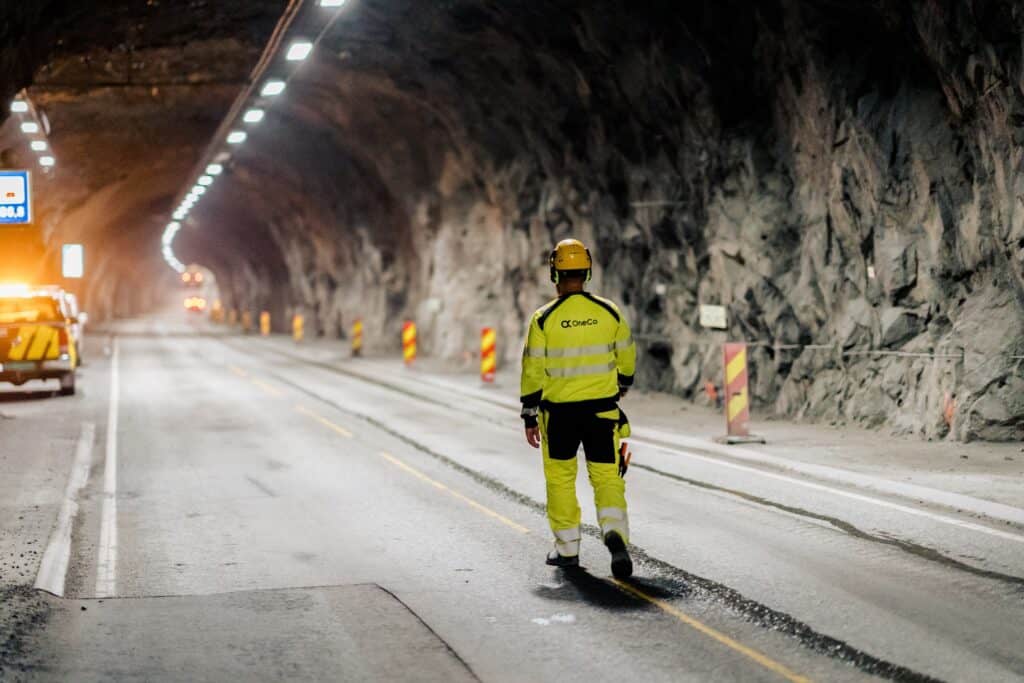 A worker is walking down the middle of a road in a tunnel away from the camera. Vehicles with lights turned on are visible in the background.