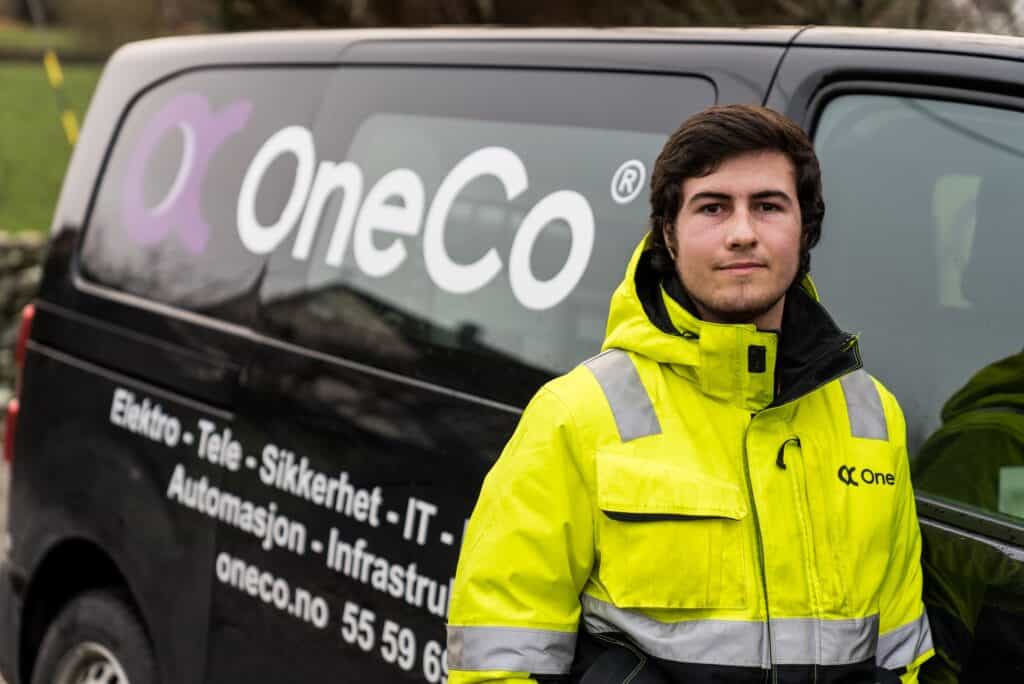 A OneCo worker is looking at the camera whilst standing beside their work car.