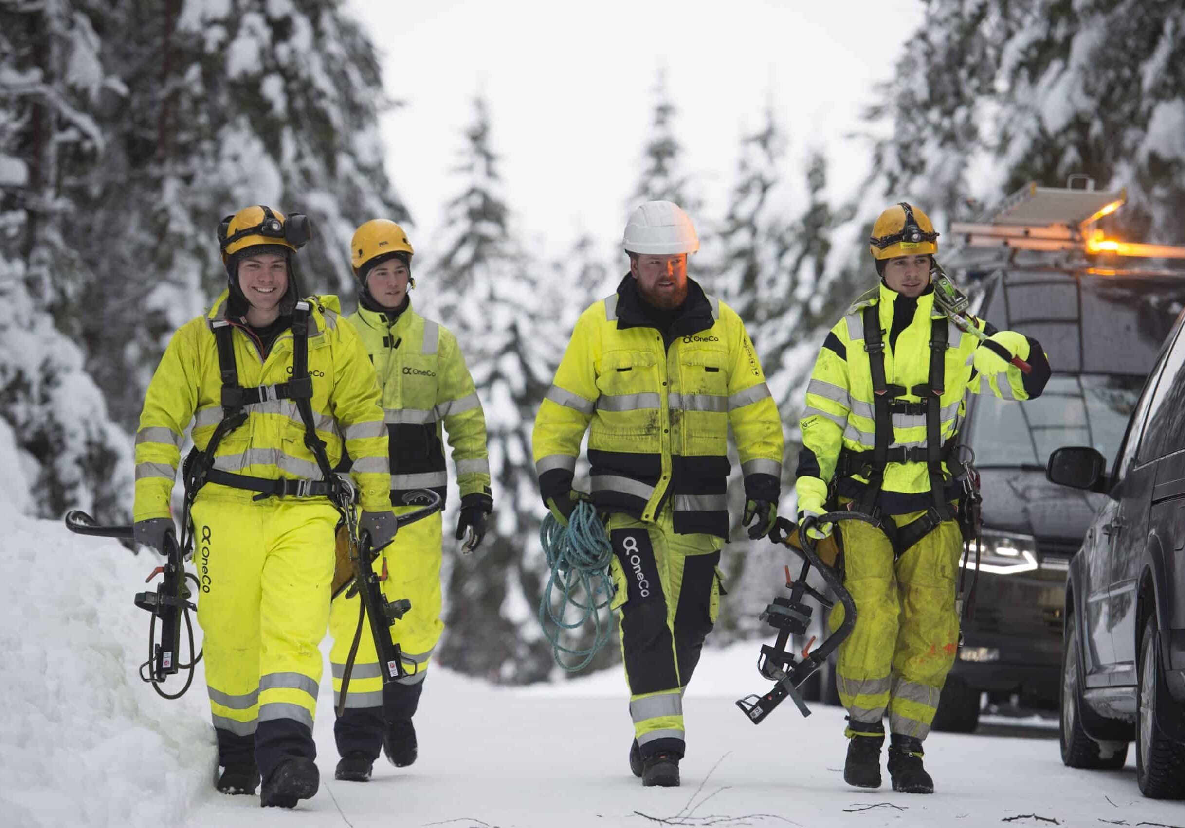El-kraft Four OneCo workers in walking through the snow in their uniforms with various equipment. Two vehicles can be seen in the background.