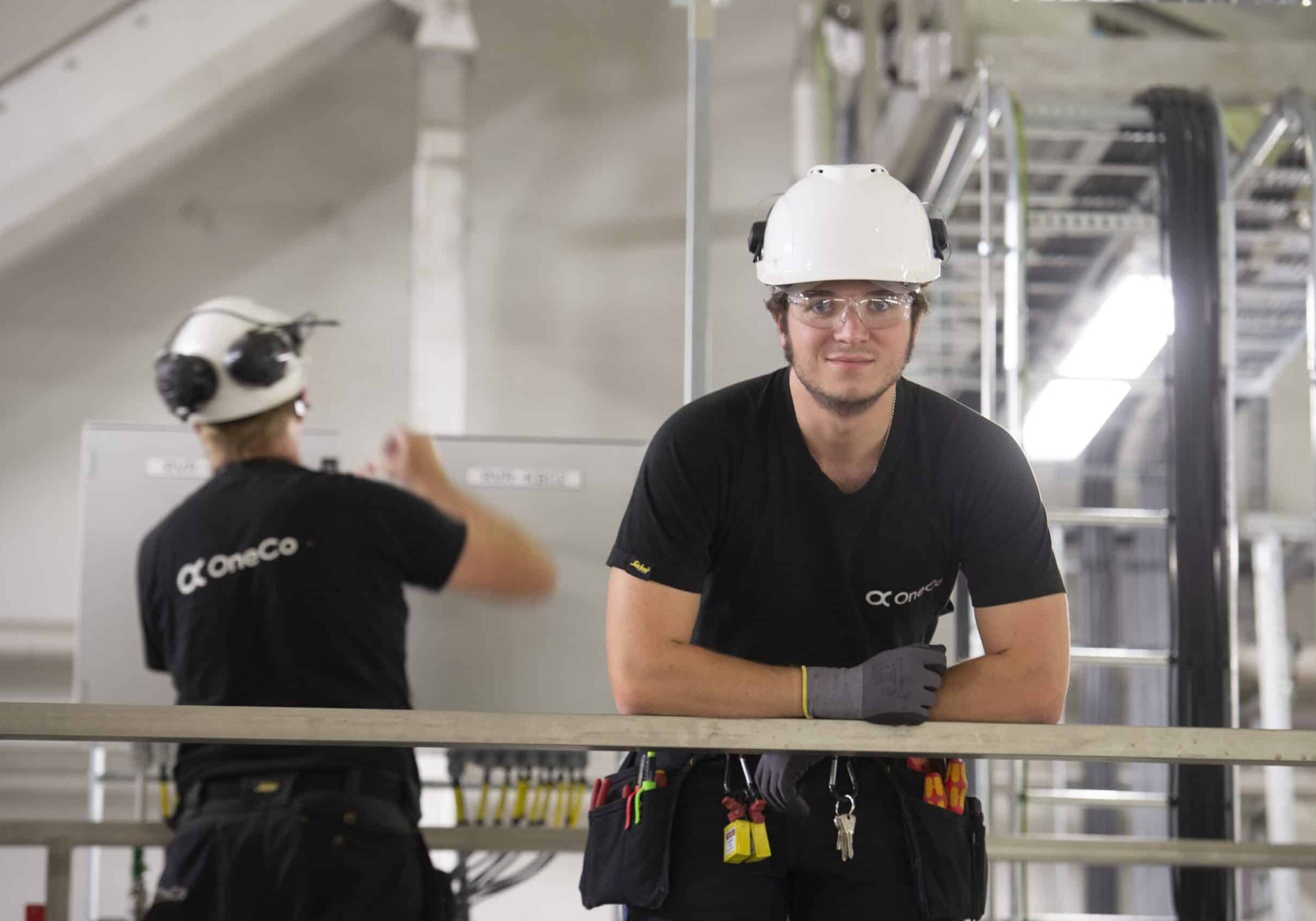 A OneCo worker is looking directly at the camera with helmet and glasses on. In the background another worker is seen fixing an electric outlet.